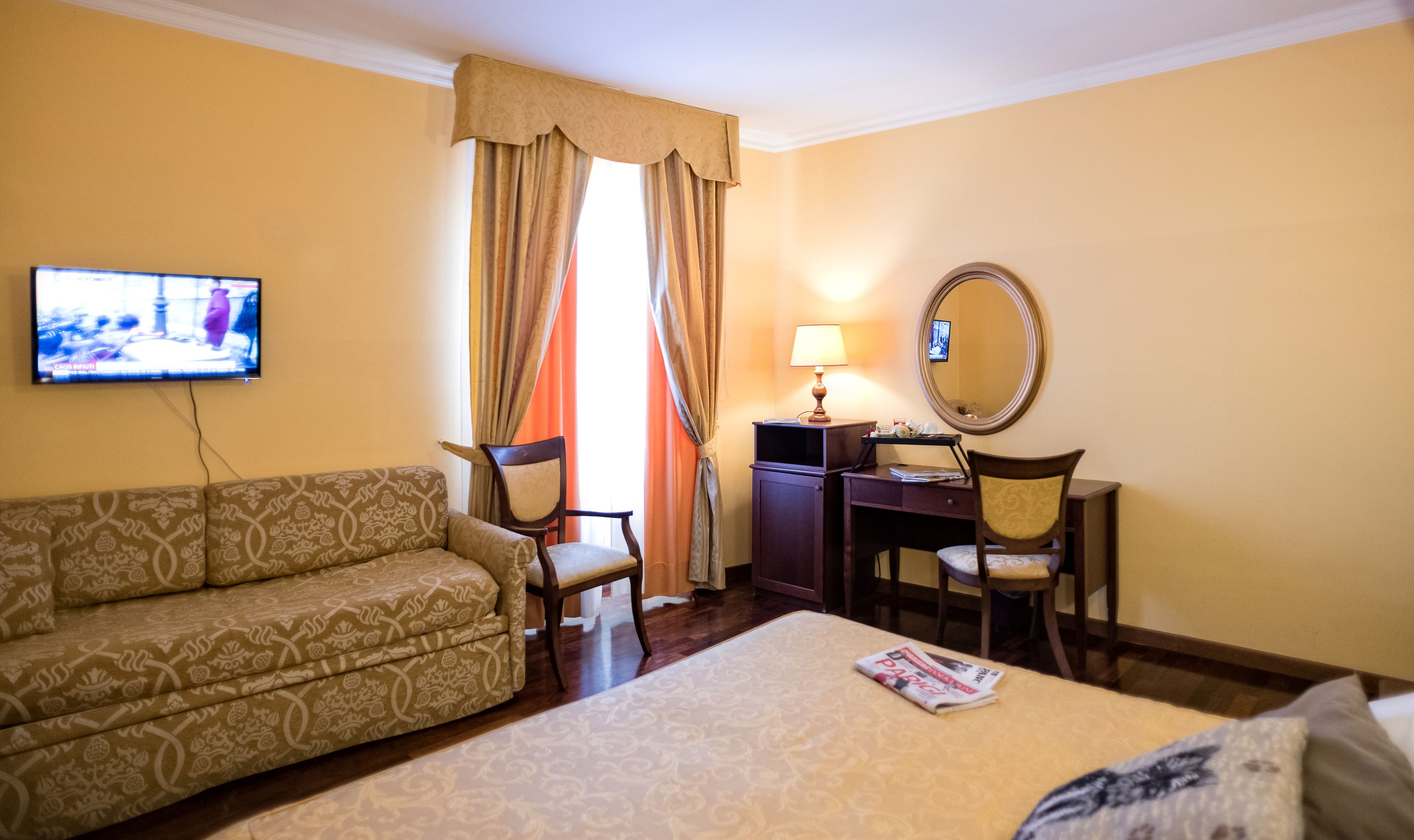 A hotel room with a sofa, a desk with a lamp, a television, and a bed.