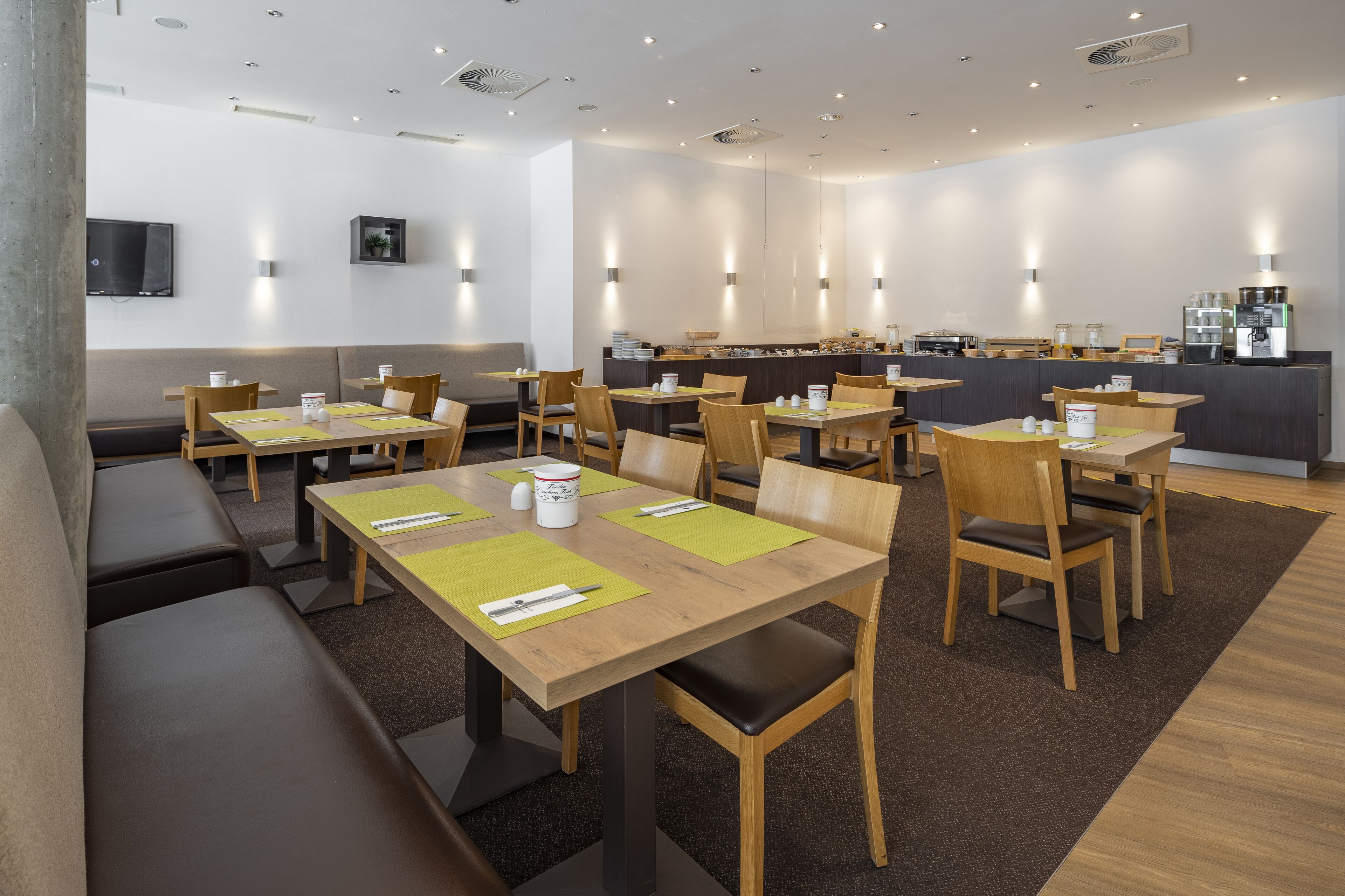 A modern restaurant with wooden tables and chairs, a counter with a coffee machine, and a TV mounted on the wall.