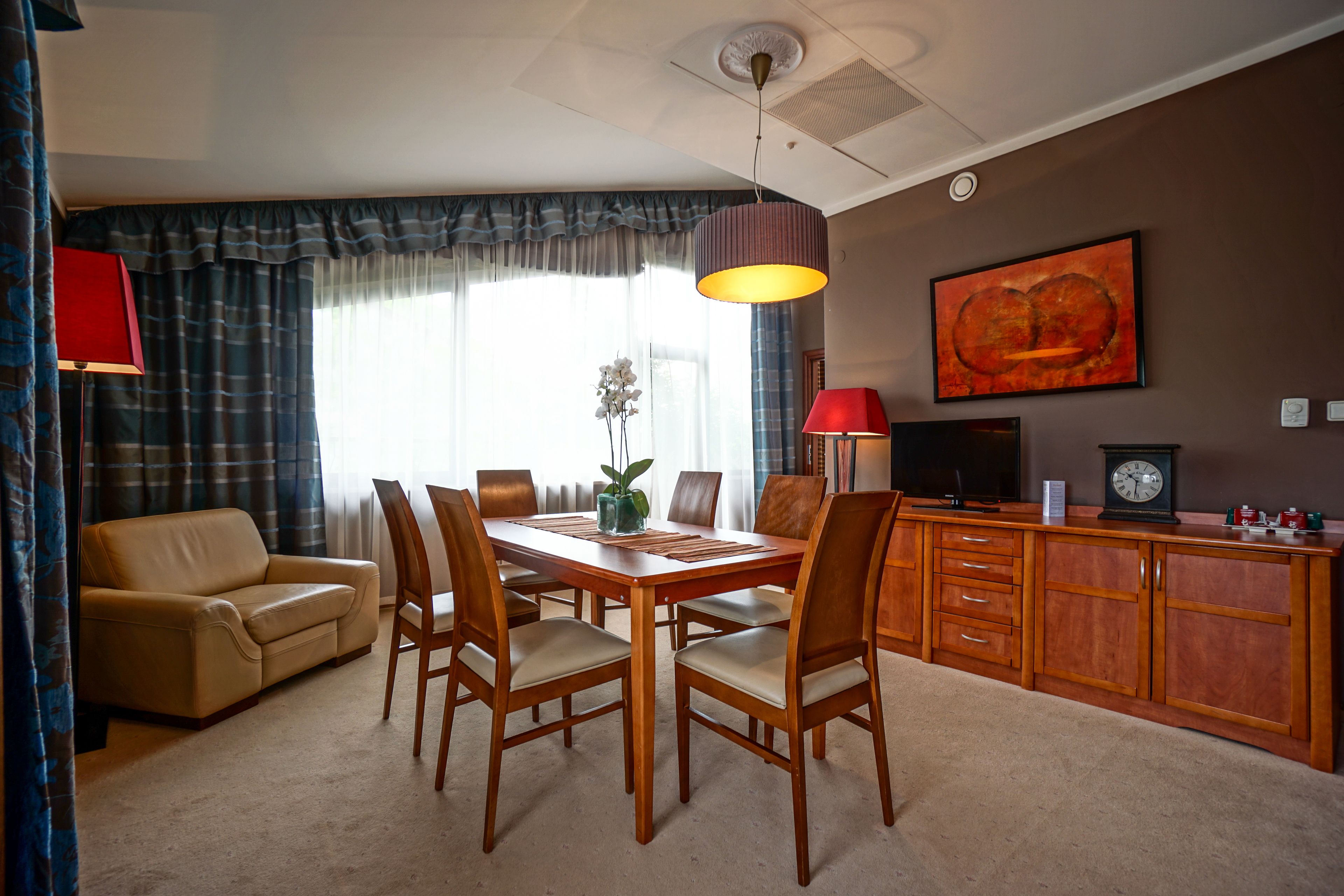 A dining area with a table, chairs, a sofa, a lamp, a clock, and a painting on the wall.