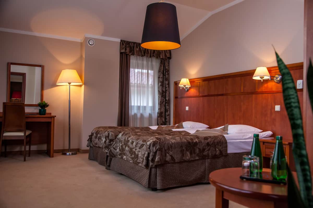Habitación de hotel con cama, escritorio, silla, lámpara y mesita de noche con botellas.