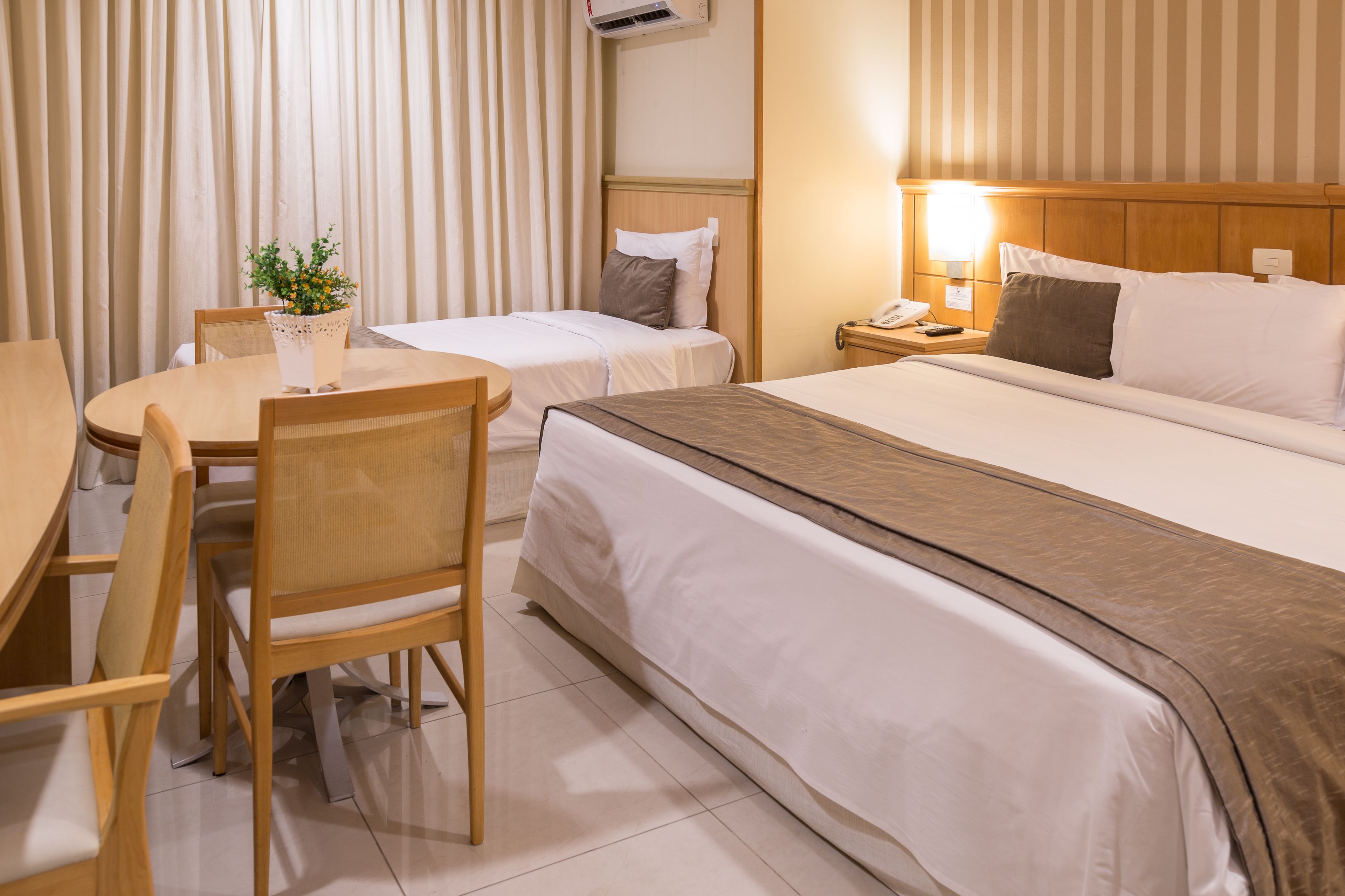 Una camera d'albergo con un letto, due comodini, una scrivania con sedie e una pianta in vaso.
