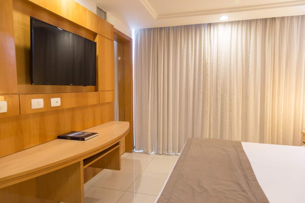 A hotel room with a wooden desk, a flat-screen TV mounted on the wall, a bed with a beige bedspread, and a large window with sheer curtains.