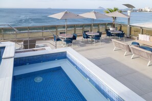 Un área de piscina en la azotea con vistas al océano, mobiliario exterior y sombrillas.