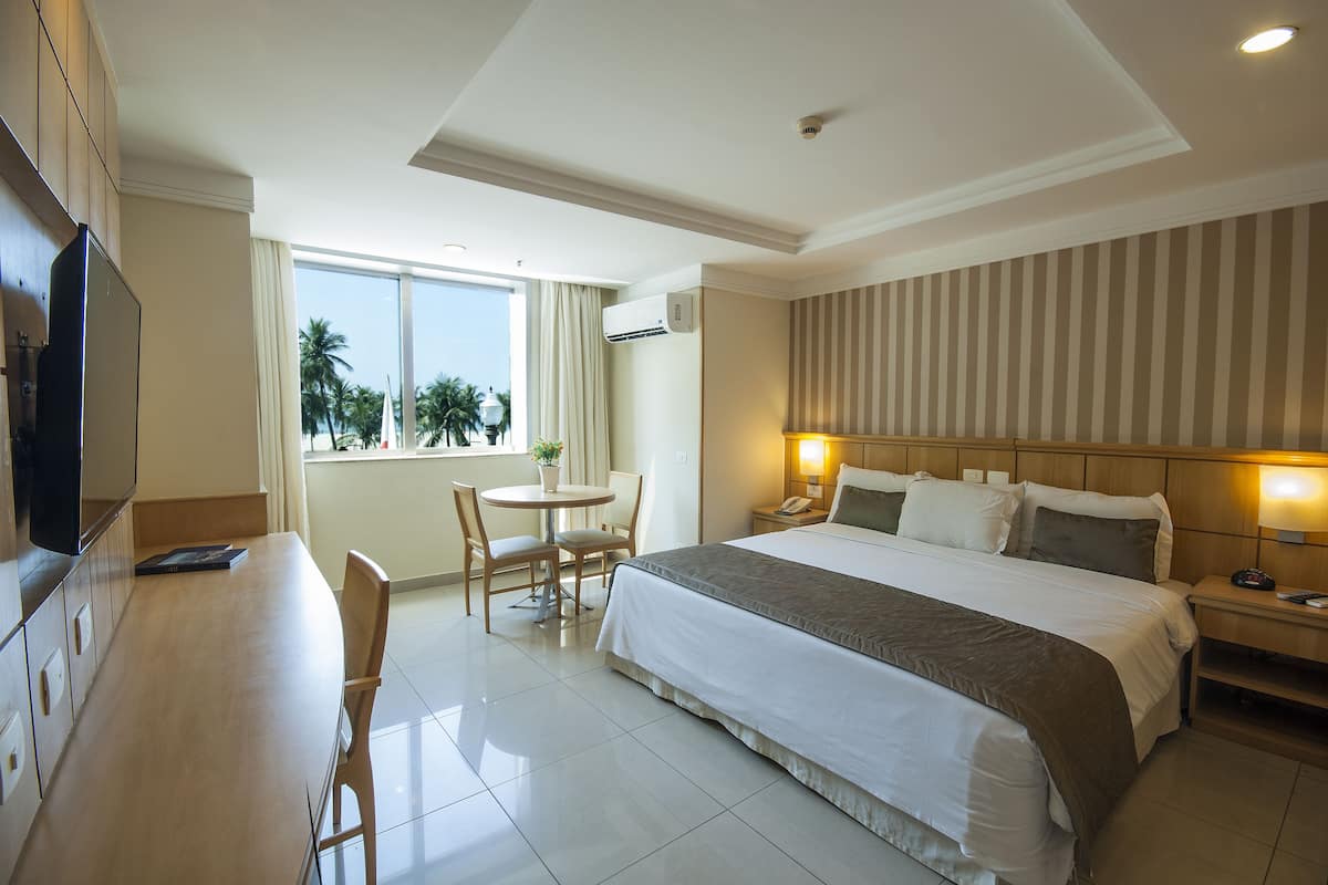 A hotel room with a bed, bedside table, lamp, TV, desk, chairs, and a window with a view of palm trees.