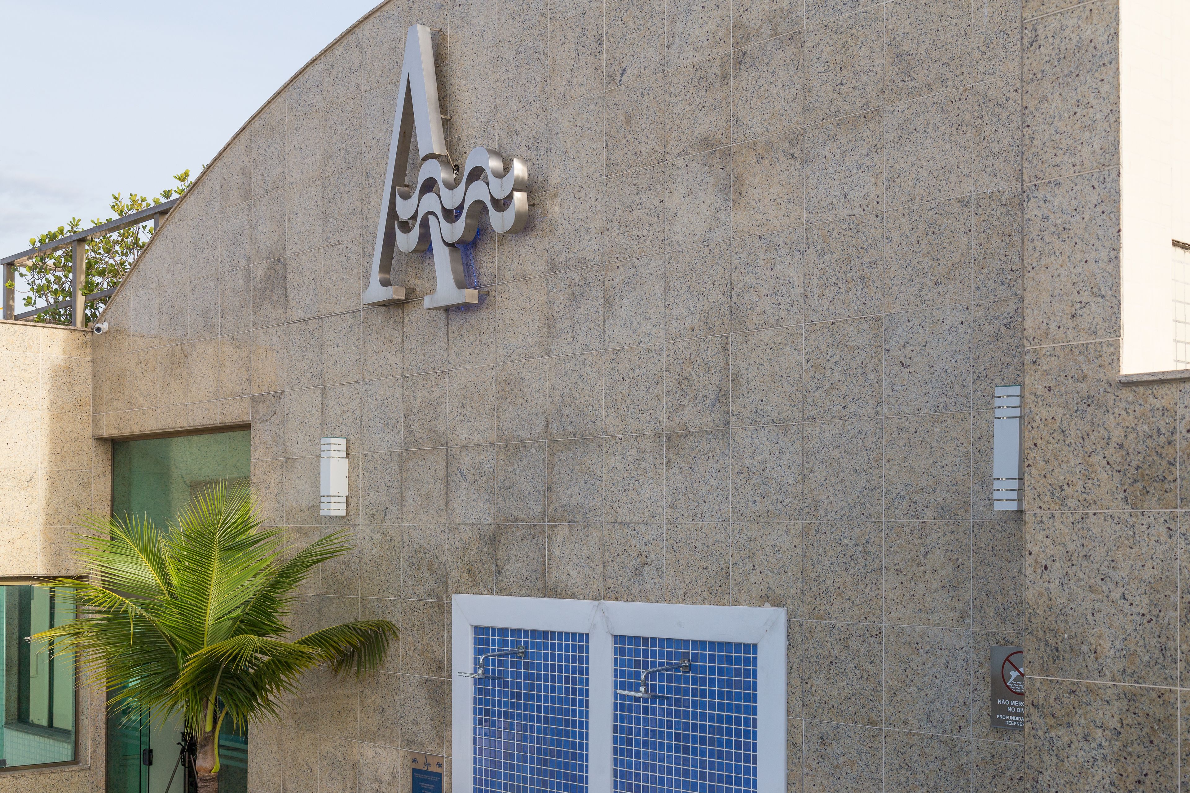 Un edificio con un gran logotipo estilizado de la letra 'A' en la fachada, una palmera en primer plano y una entrada con azulejos azules.