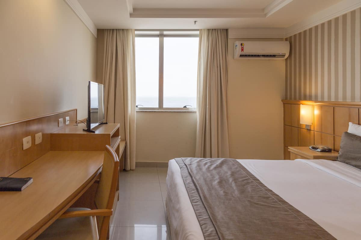 A hotel room with a bed, a desk, a chair, a TV, a window with curtains, an air conditioner, and a nightstand with a lamp.