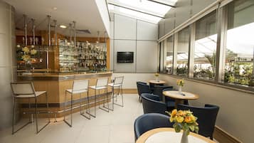 A modern bar area with a high ceiling, glass walls, and a view of greenery outside.