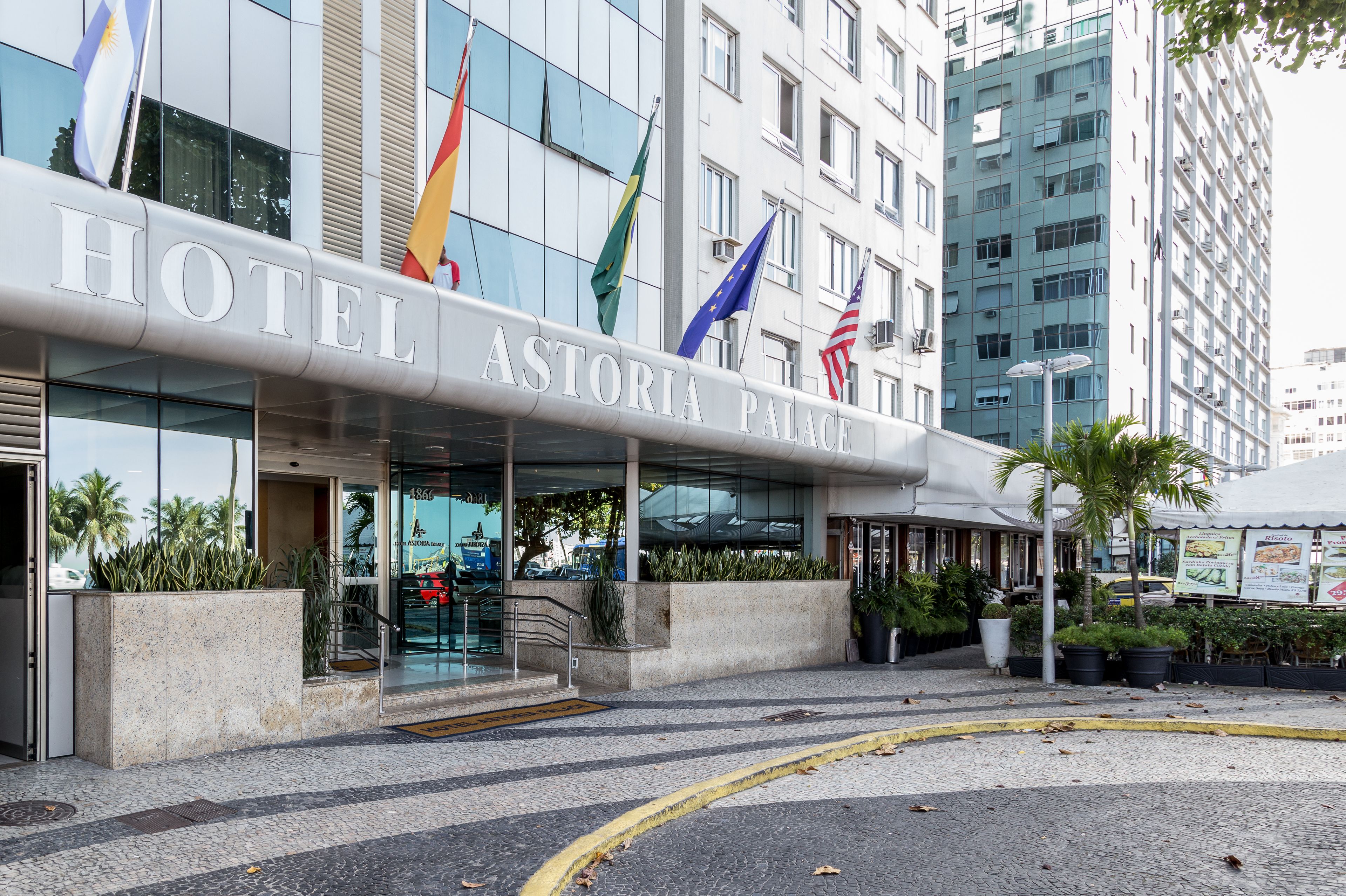 A hotel entrance with a modern glass facade, flags, and a paved area with plants.
