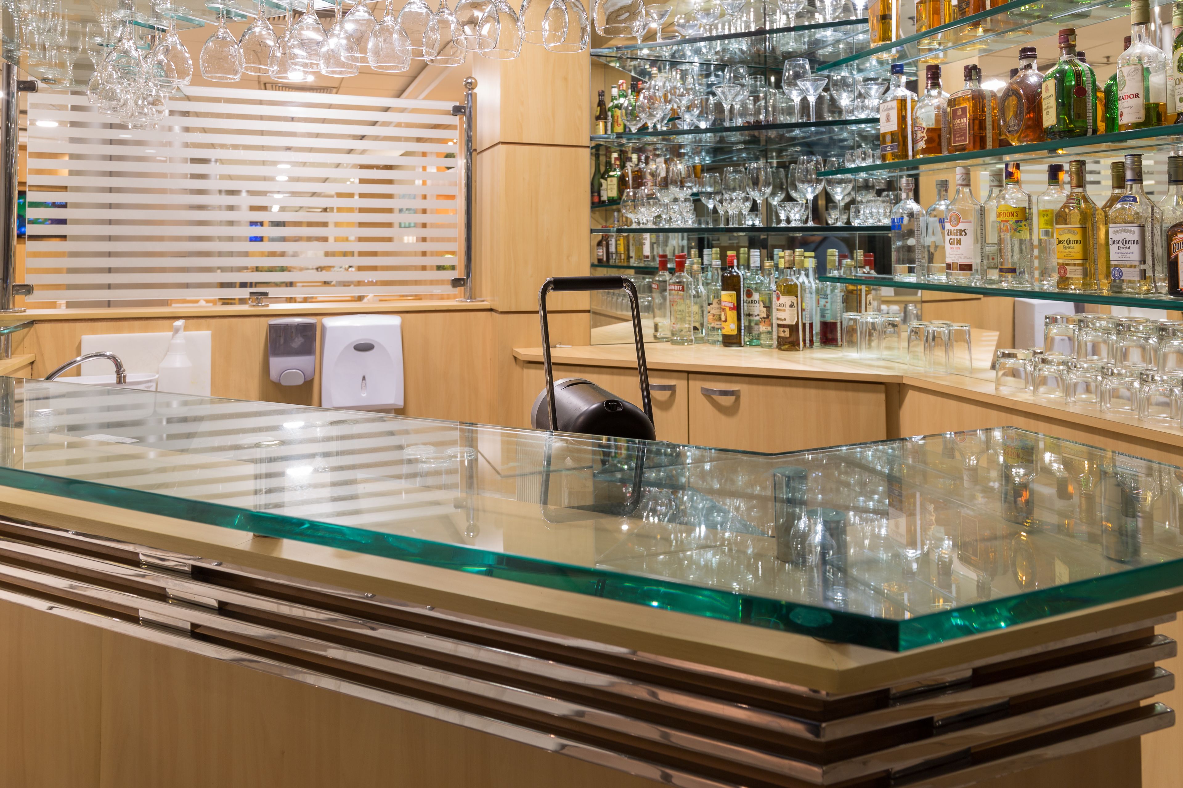 Un espace bar doté d’un comptoir en verre, d’étagères remplies de bouteilles d’alcool et de verres à vin suspendus.