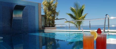 Vista de la piscina con dos bebidas, una palmera y cielo despejado.