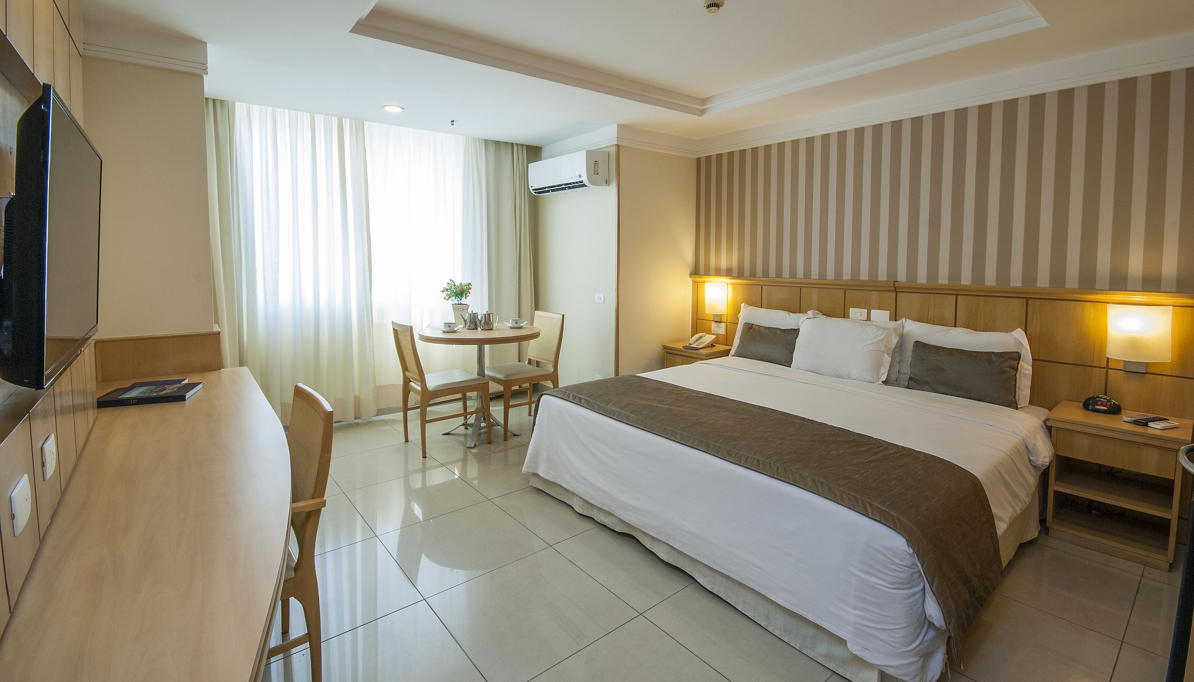 A hotel room with a bed, bedside table, lamp, chair, desk, TV, and an air conditioning unit.