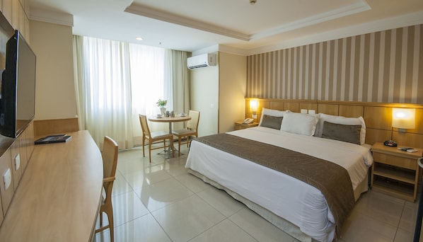 Habitación de hotel con cama, mesita de noche, lámpara, silla, escritorio, televisión y aire acondicionado.