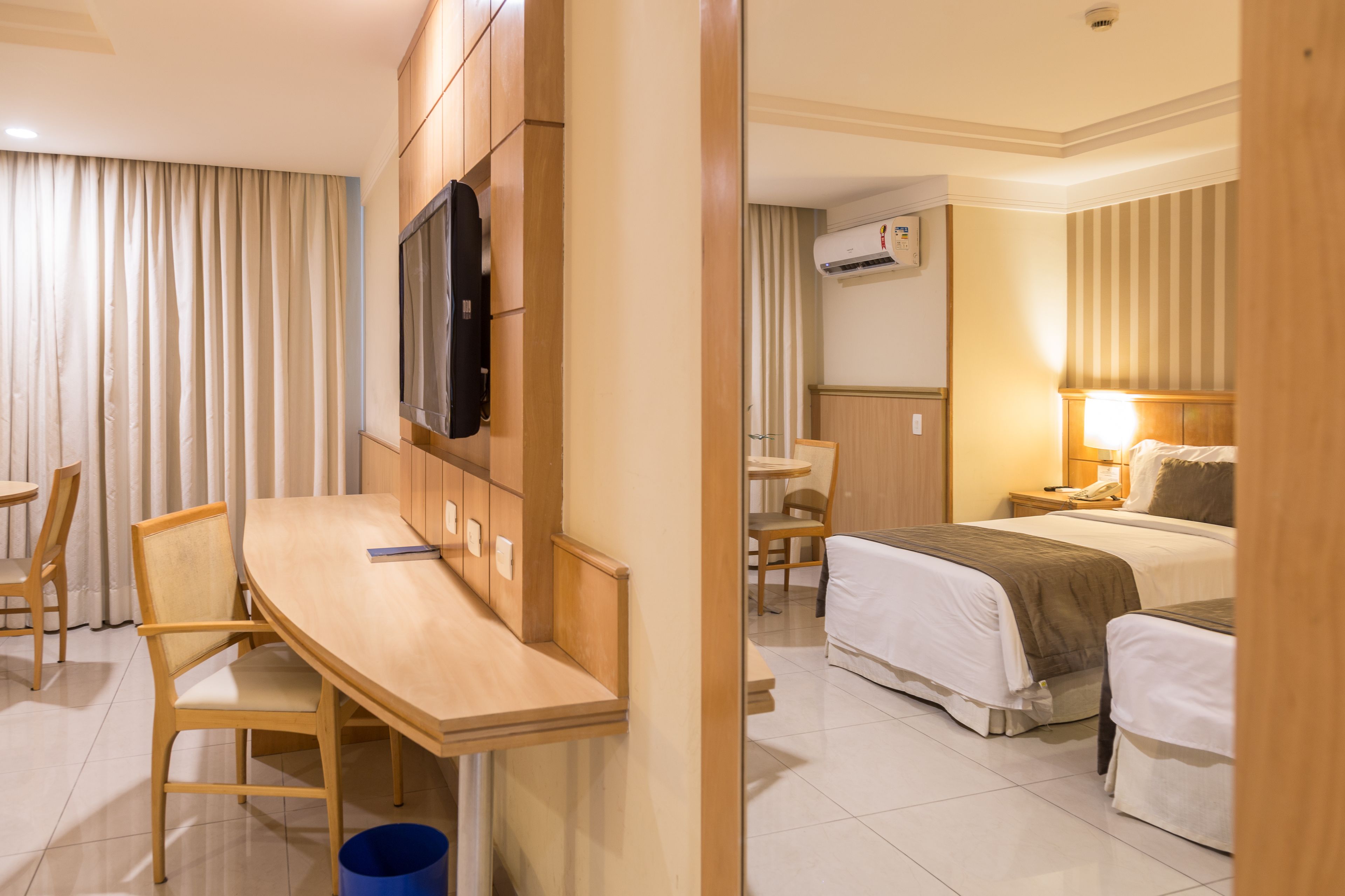 A hotel room with a wooden desk, two beds, a TV, and an air conditioning unit.