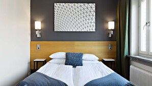 Ein ordentlich bezogenes Bett mit blauer Tagesdecke, zwei Nachttischen, einer Wandleuchte und einem Fenster mit Vorhängen.