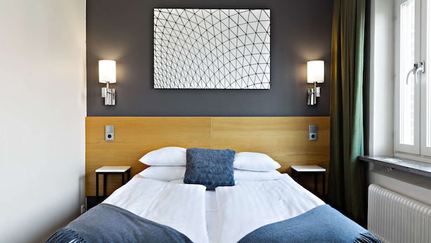 Een net opgemaakt bed met een blauwe deken, twee nachtkastjes, een wandlamp en een raam met gordijnen.