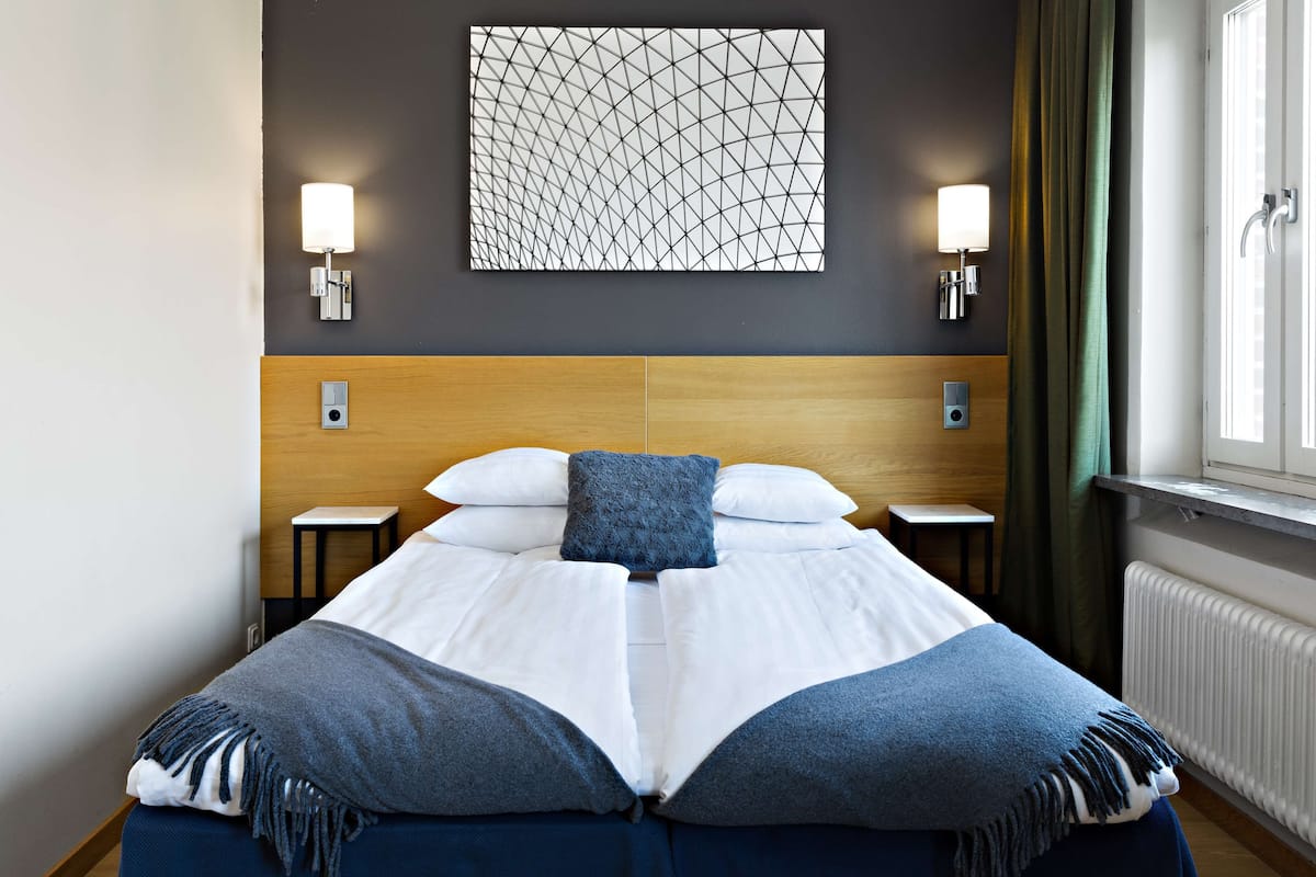 Un lit bien fait, recouvert d’un plaid bleu, deux tables de chevet, un luminaire fixé au mur et une fenêtre avec des rideaux.