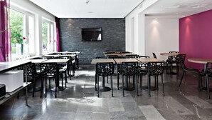 Een moderne eetruimte met meerdere tafels en stoelen, een flatscreen televisie bevestigd aan een bakstenen muur, en grote ramen die natuurlijk licht binnenlaten.