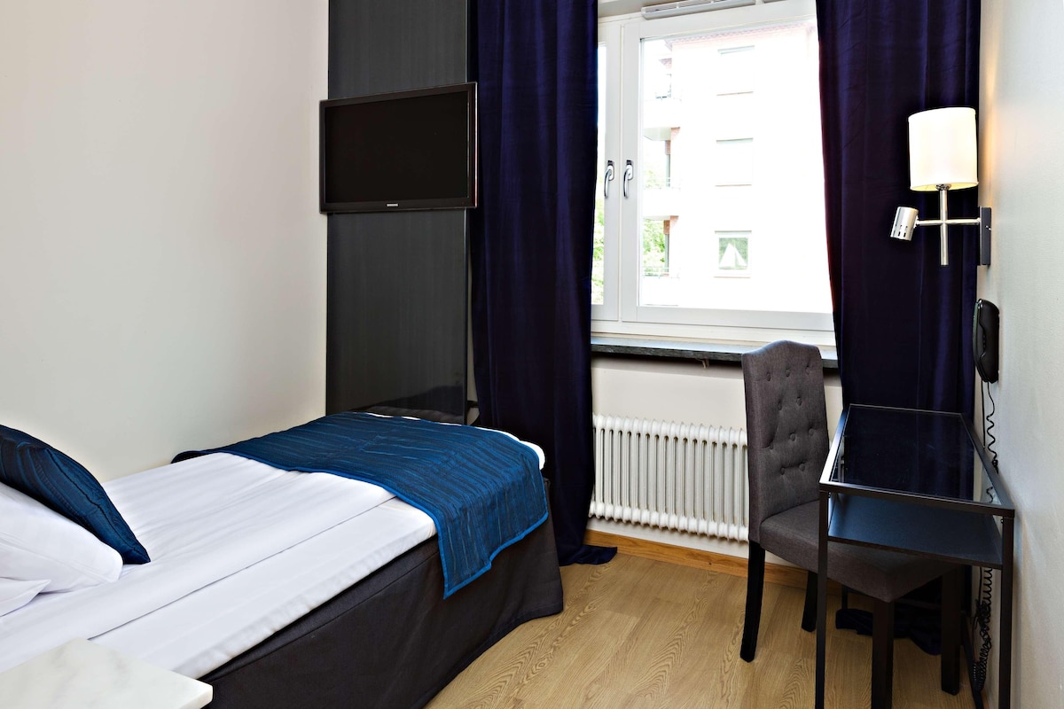 Een hotelkamer met een bed, een bureau, een stoel, een aan de muur bevestigde televisie en een raam met gordijnen.