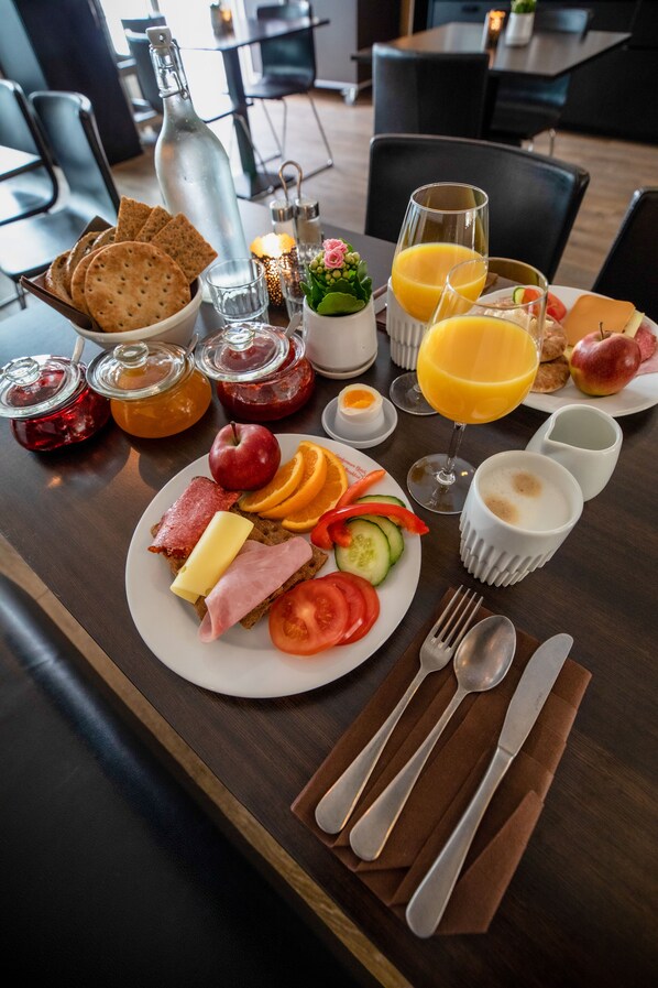 Una escena de desayuno con una fuente de tostadas, jamón, queso y verduras, acompañada de un vaso de zumo de naranja, una botella y varios condimentos.