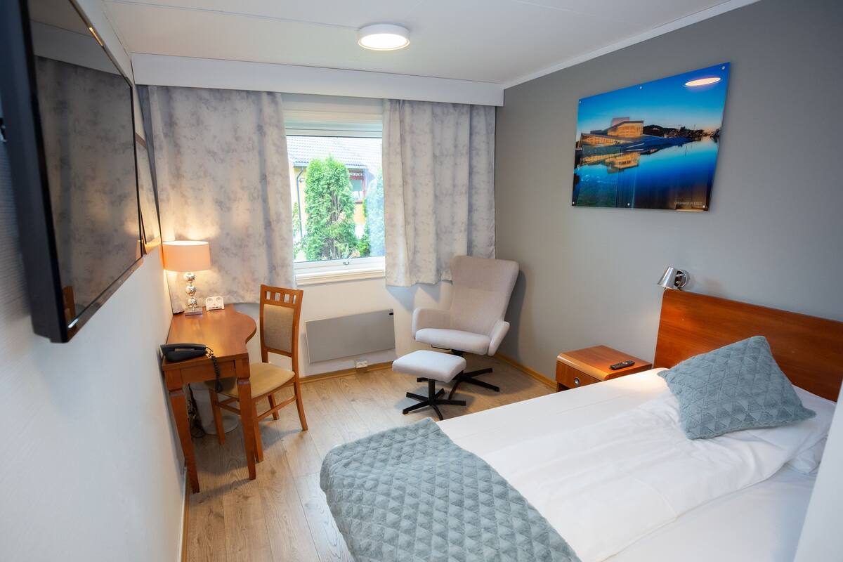 A hotel room with a bed, a desk with a chair, a window with curtains, a nightstand, and a painting on the wall.