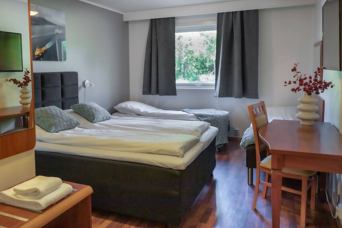 A hotel room with two beds, a wooden desk, a chair, a TV, and a window with curtains.