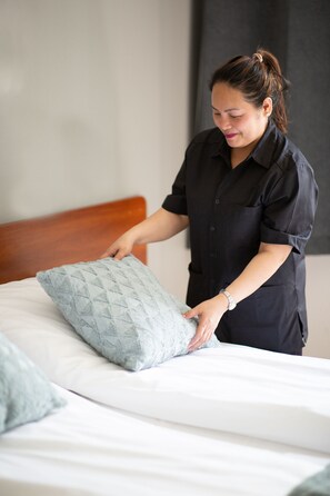 A person adjusting a pillow on a bed with a wooden headboard in a room with a plain wall. - Gardermoen Hotel Bed & Breakfast (Nannestad)