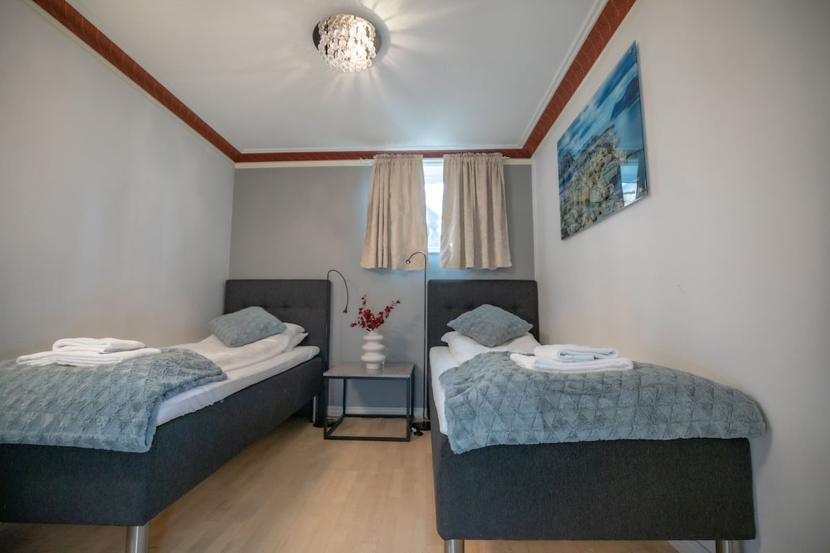 A room with two beds, a small table with a vase, and a painting on the wall.