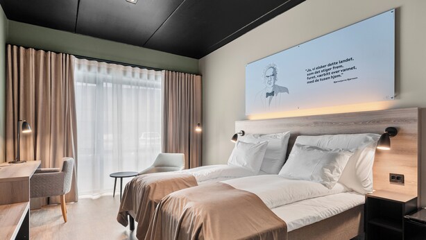 A modern hotel room with a large bed, a desk, and a chair. There is a window with curtains and a wall-mounted artwork. - Hotell Jæren (Time)