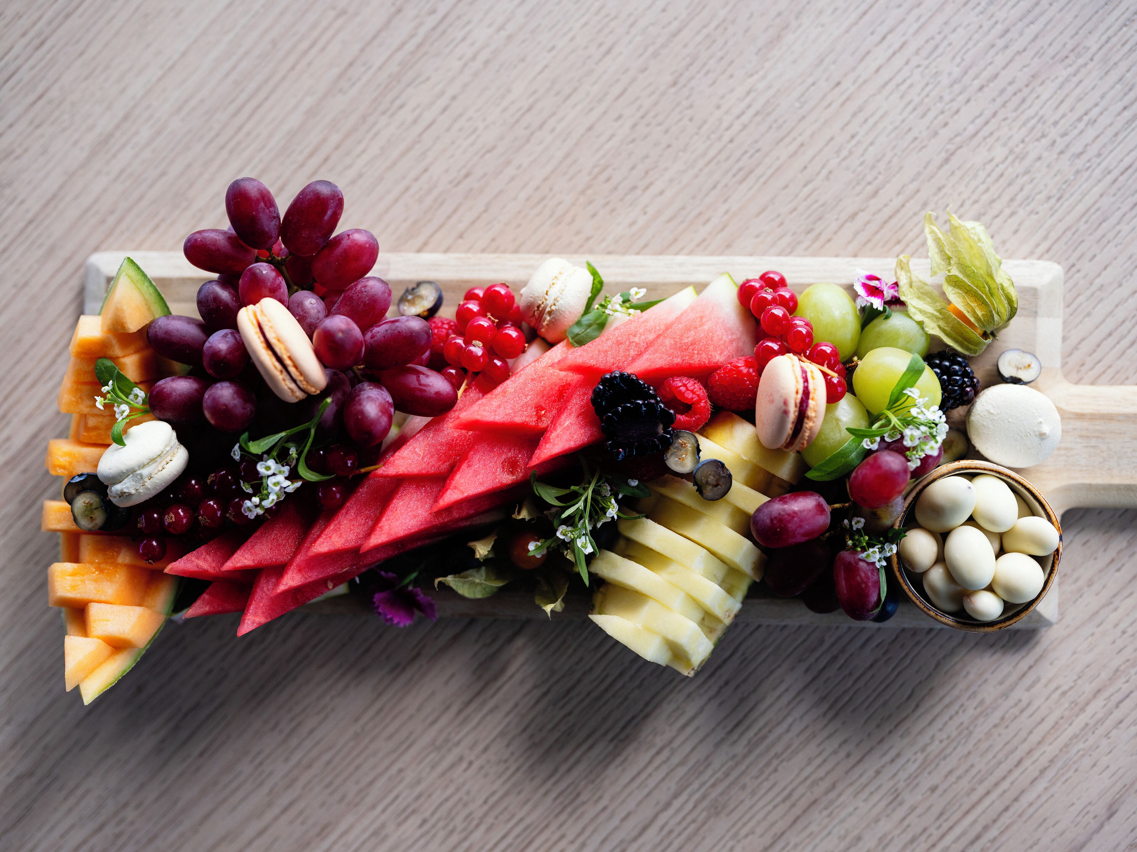 Una tabla de madera con una variedad de frutas, como uvas, sandía y manzanas, además de algunos pasteles y un recipiente pequeño con huevos.
