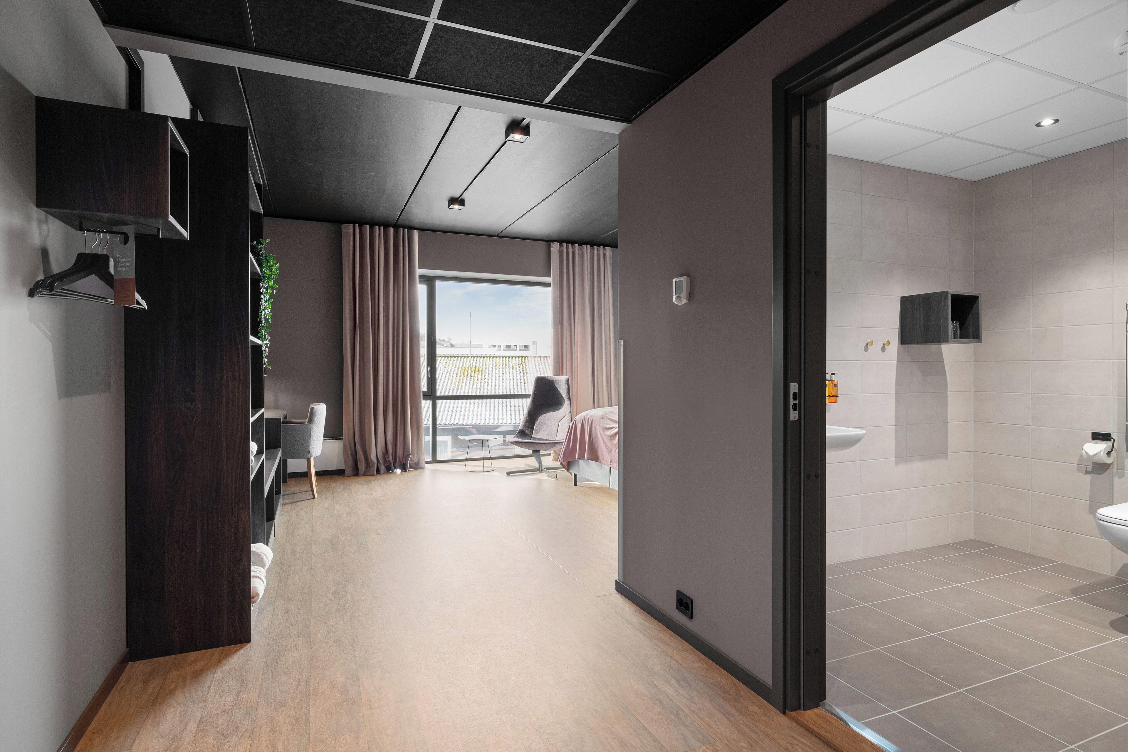 Una habitación de hotel moderna con piso de madera, un balcón con vistas, un baño con inodoro y lavamanos, y un escritorio con silla.