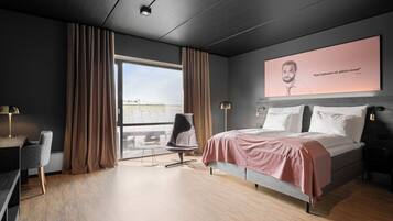 A modern hotel room with a large bed, a desk, a chair, and a view of the city through the window.