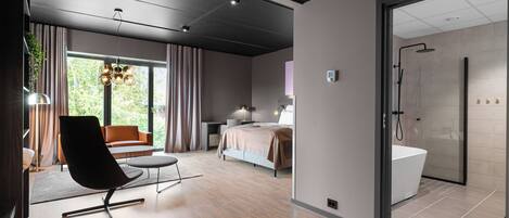 A modern hotel room with a large bed, a sitting area with a chair and a table, a shower area, and a view of greenery outside.