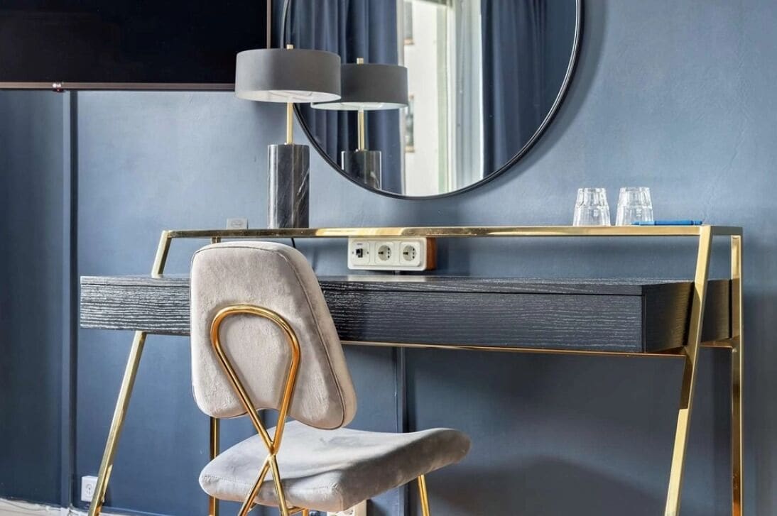 Un tocador moderno con marco dorado, un espejo redondo y una silla gris.