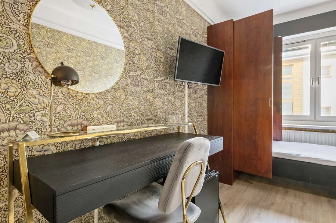 A room with a dark wooden desk, a round mirror, a flat-screen TV, and a patterned wallpaper.