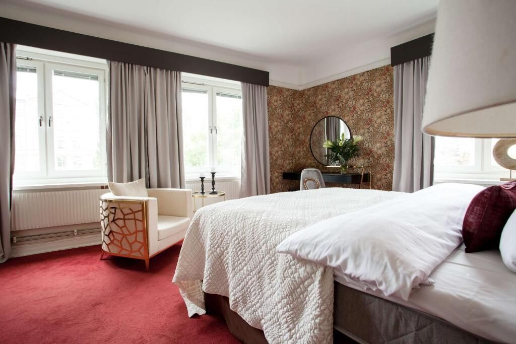 Ett hotellrum med en stor säng, en stol, en rund spegel och ett fönster med gardiner.