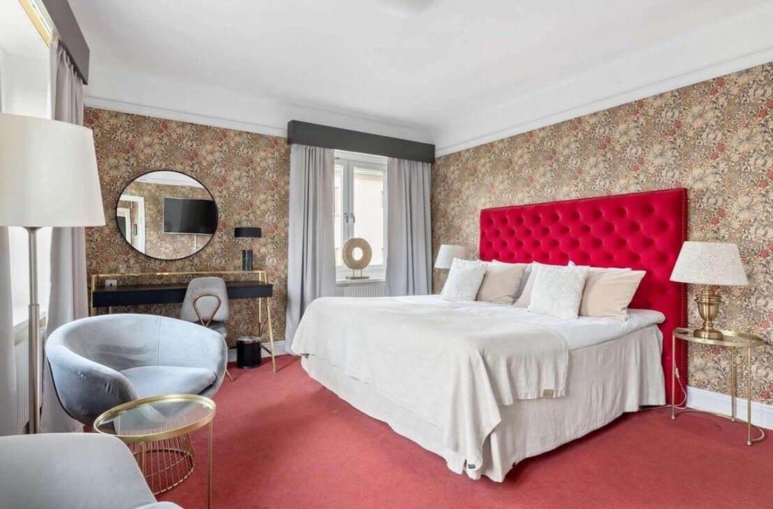 Een hotelkamer met een groot bed, een rood hoofdeinde, een ronde spiegel, een bureau met een televisie en een stoel.