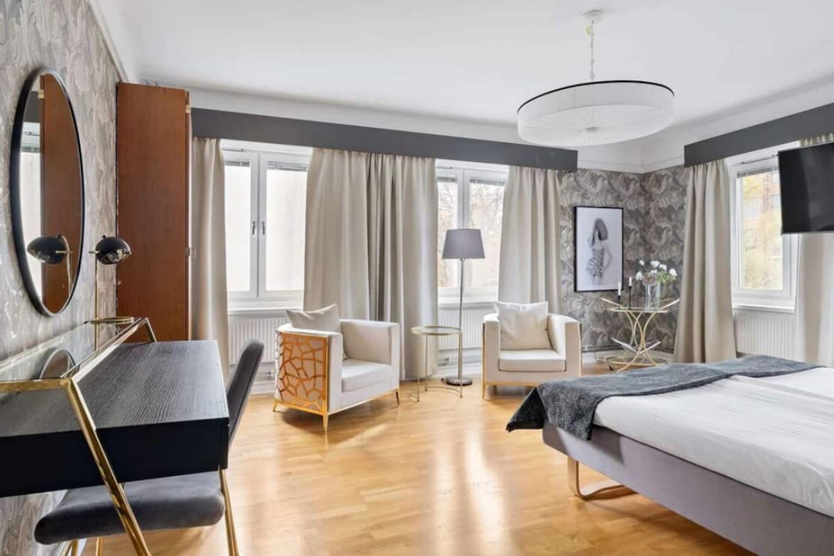 Une chambre d’hôtel moderne avec un plancher en bois, un lit, un bureau avec une chaise, deux fauteuils, une petite table et un grand miroir.