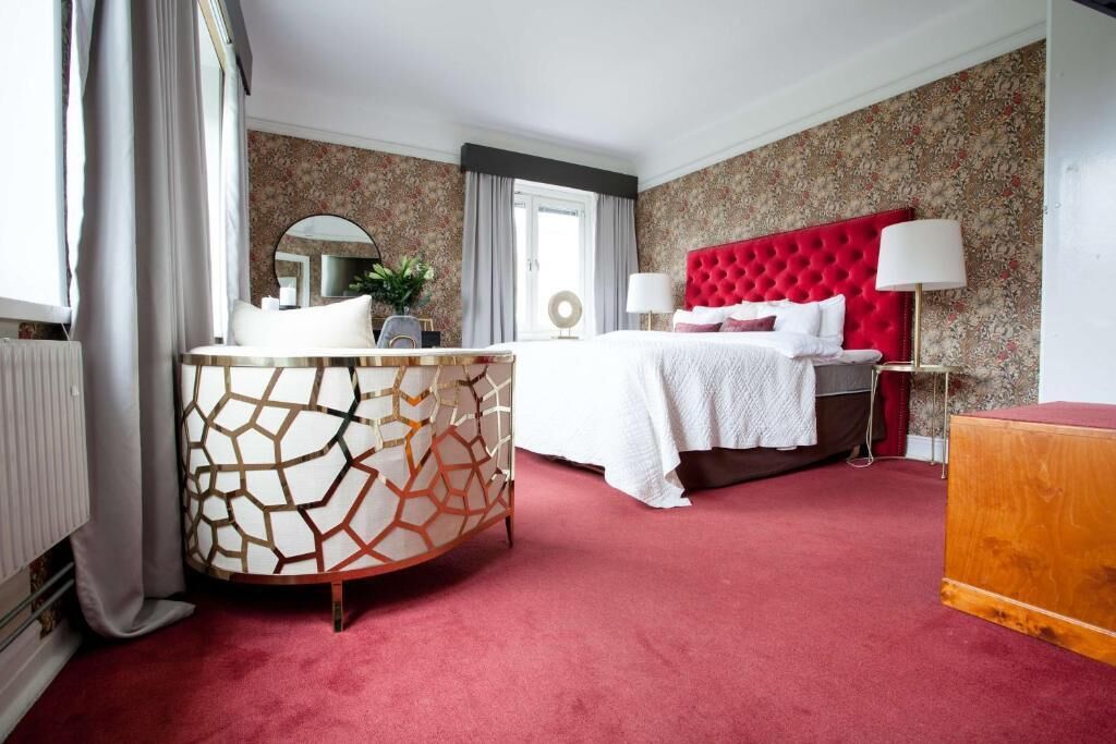Ett hotellrum med en röd, klädd sänggavel, ett runt sidobord med ett geometriskt mönster, en säng med vita sängkläder och en röd matta.