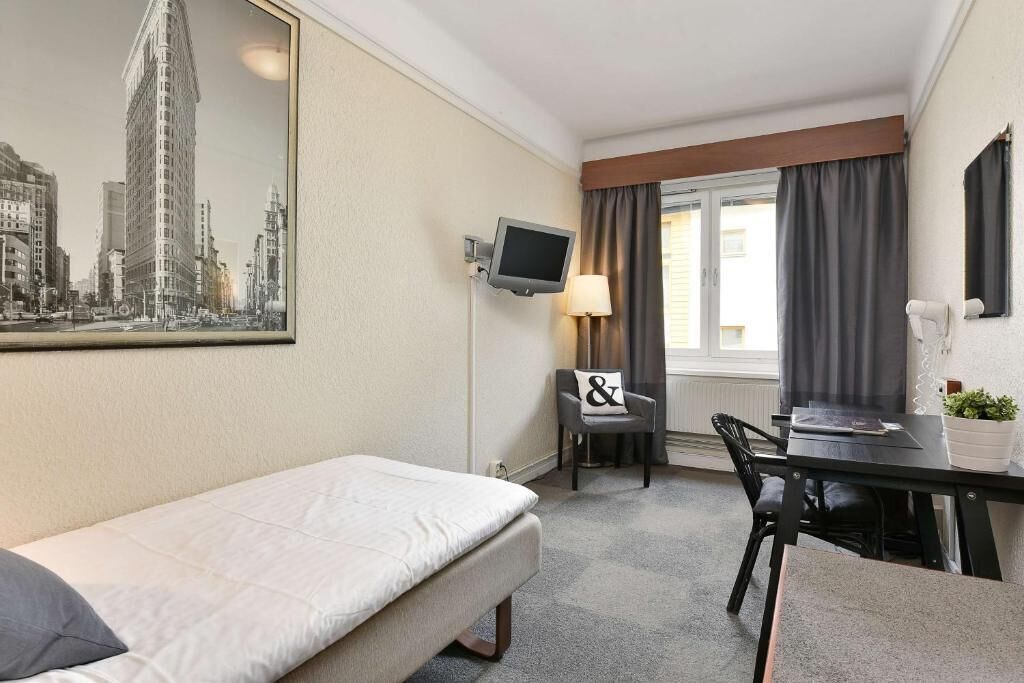 Een hotelkamer met een bed, een bureau, een stoel, een televisie, een raam met gordijnen en een stadsgezichtschilderij aan de muur.