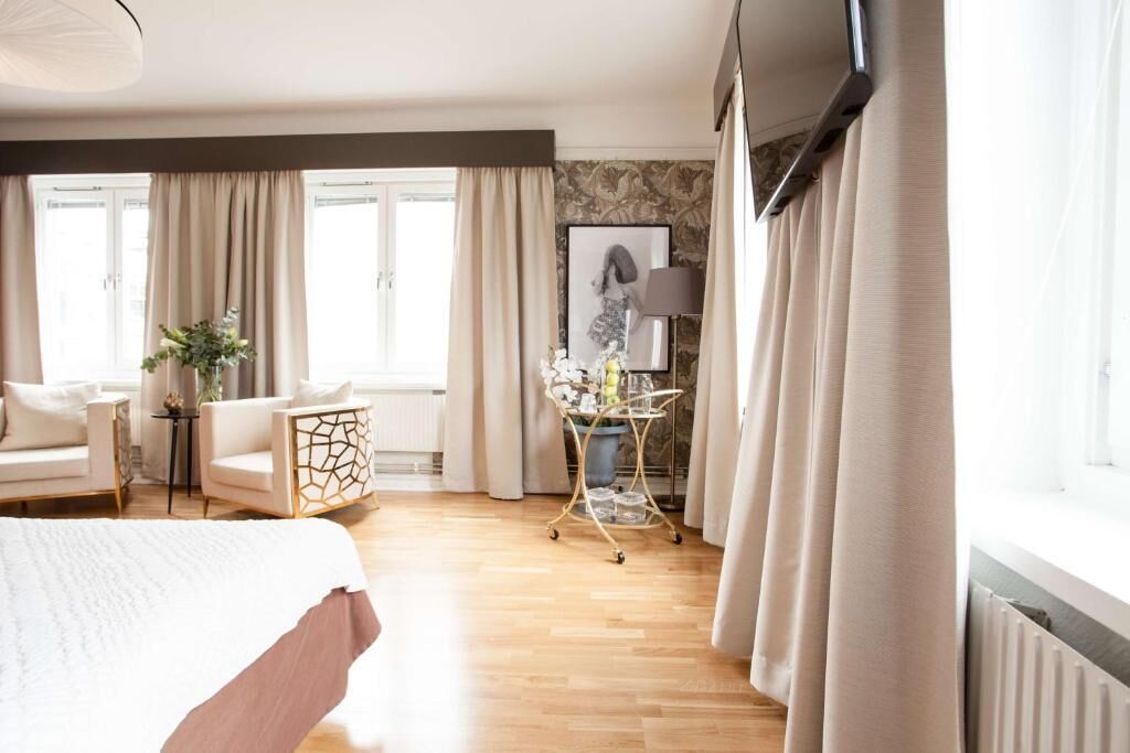 A hotel room with a bed, a sitting area with a chair and a table, a window with curtains, and a radiator.