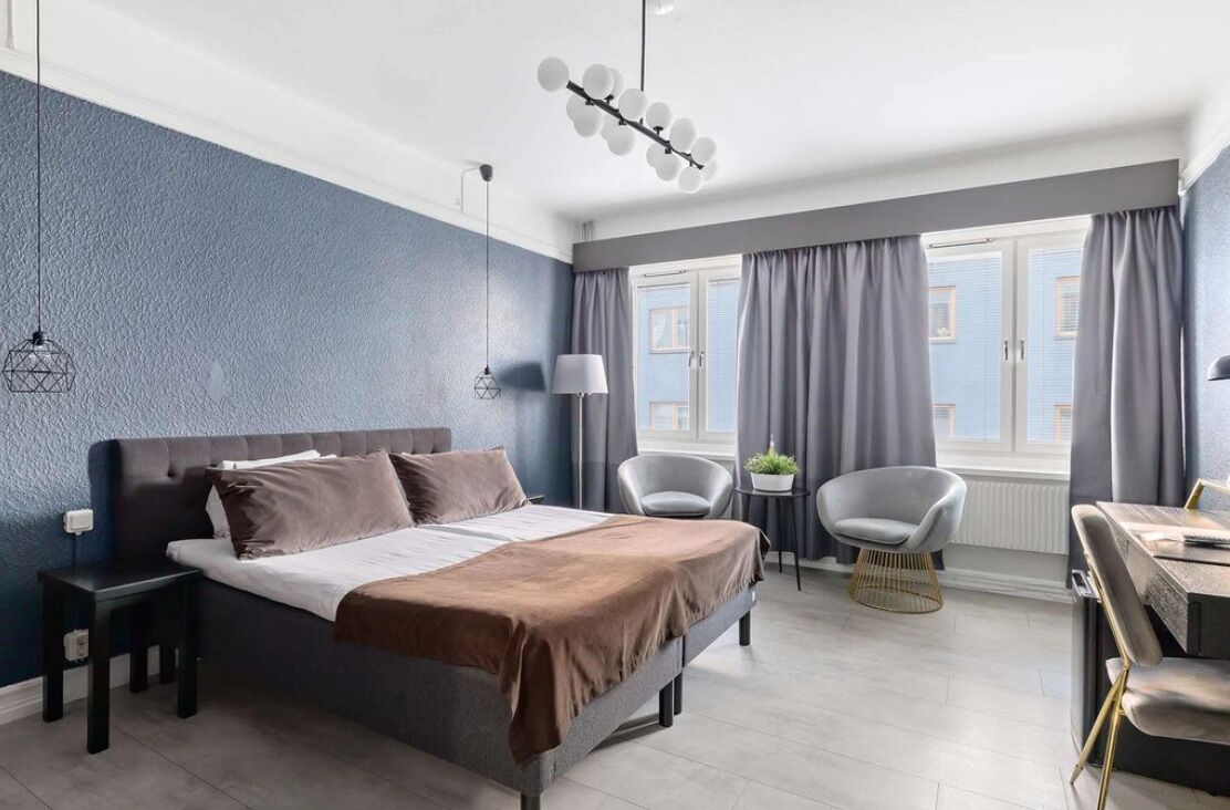 Habitación de hotel moderna con una cama grande, un escritorio y una silla. Cuenta con una ventana con cortinas, una lámpara y una lámpara colgante.