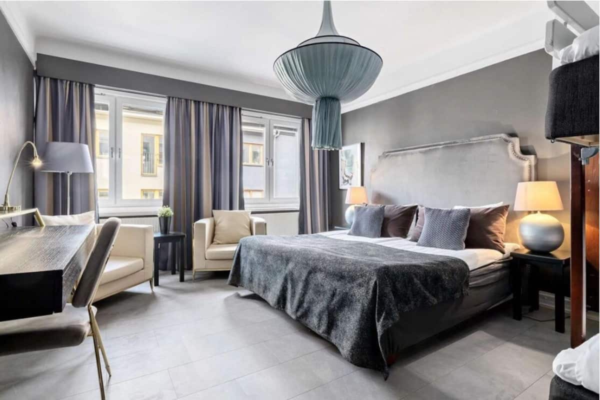 Une chambre d’hôtel moderne avec un grand lit, un bureau et une chaise. On y trouve une fenêtre avec des rideaux, un luminaire suspendu et une table de chevet avec une lampe.