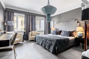 Moderni hotellihuone, jossa on suuri sänky, työpöytä ja tuoli. Huoneessa on ikkuna, jossa on verhot, riippuvalaisin ja yöpöytä, jossa on lamppu.