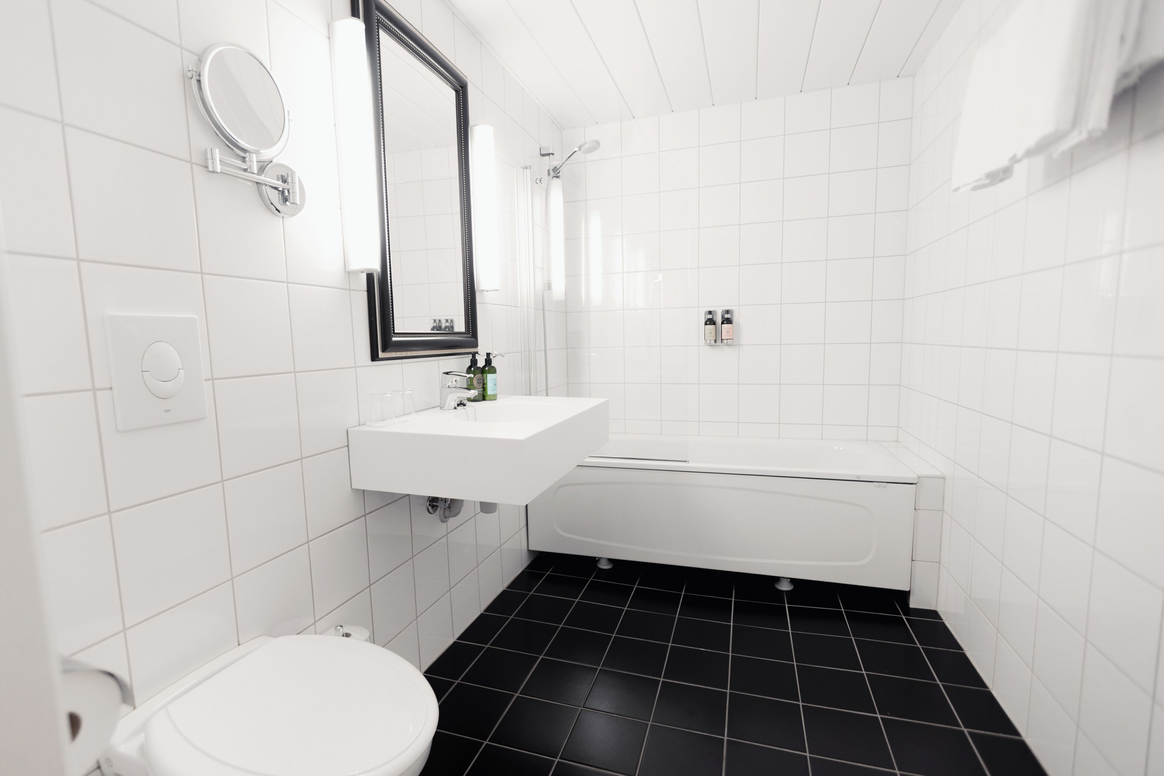 Un baño con lavamanos, espejo e inodoro blancos, suelo de baldosas negras y bañera blanca.
