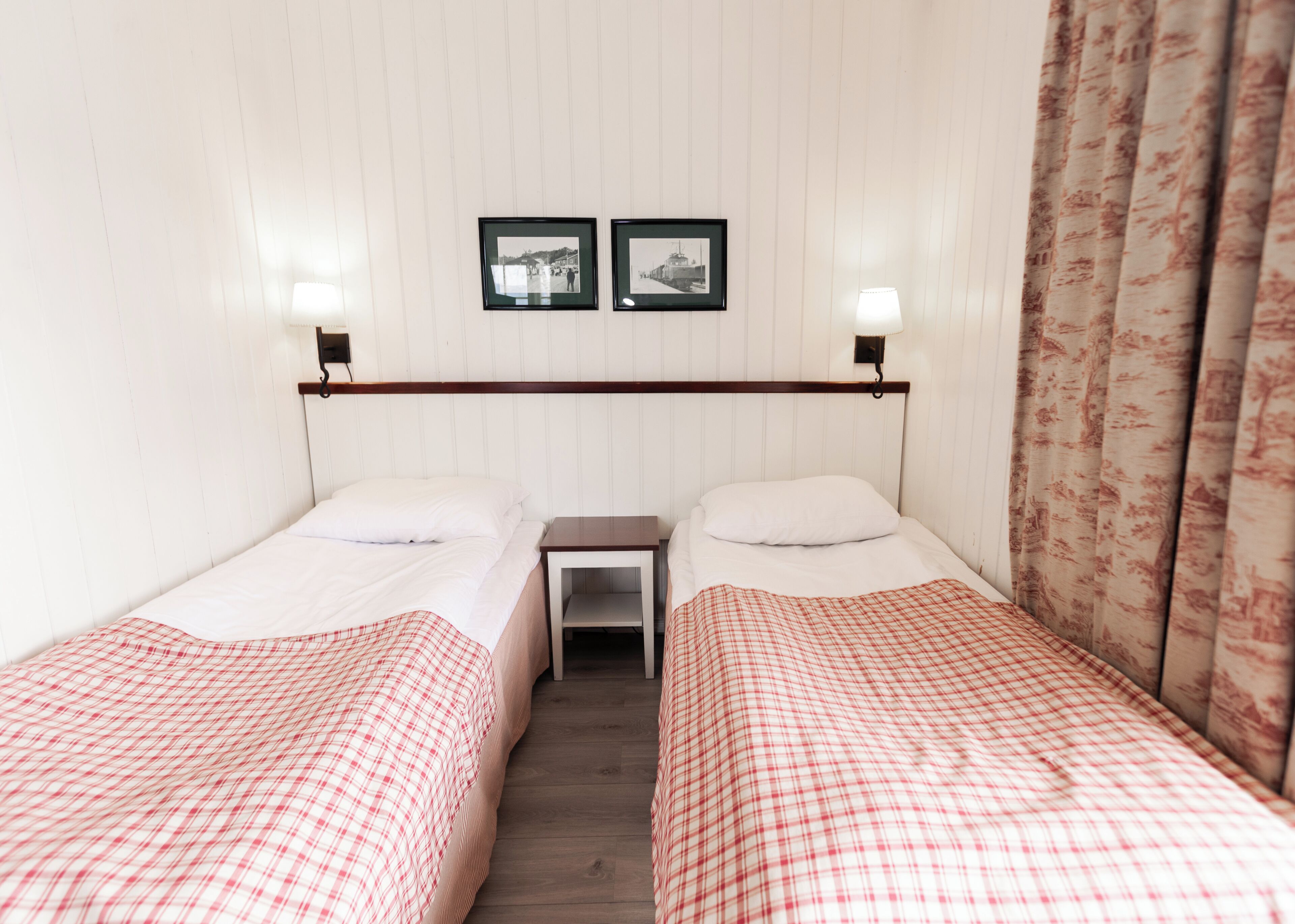 A room with two beds, each with a red and white checkered bedspread, a small bedside table, and a wall-mounted shelf above.