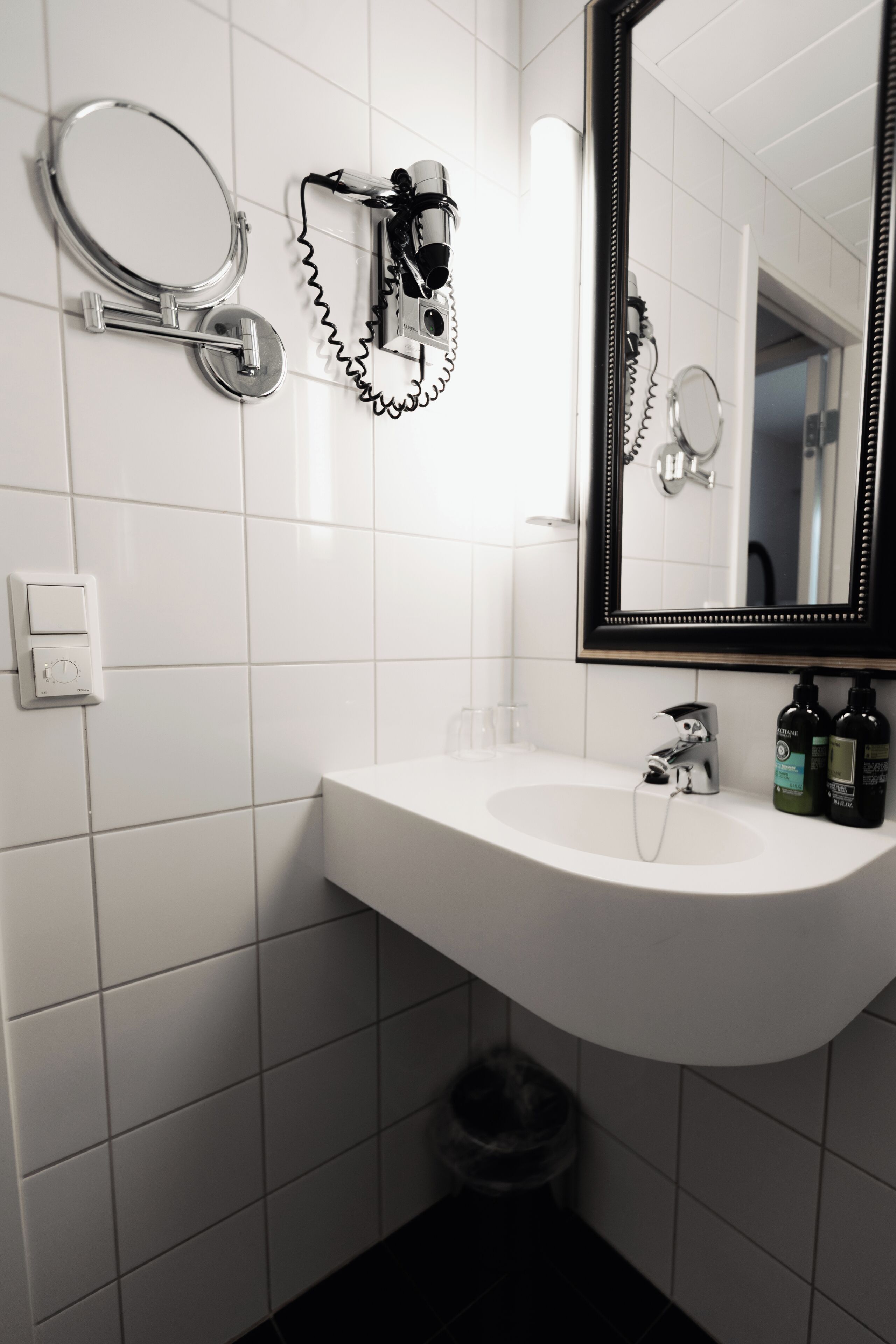 Een badkamer met een witte wastafel, een spiegel, een haardroger en een prullenbak.