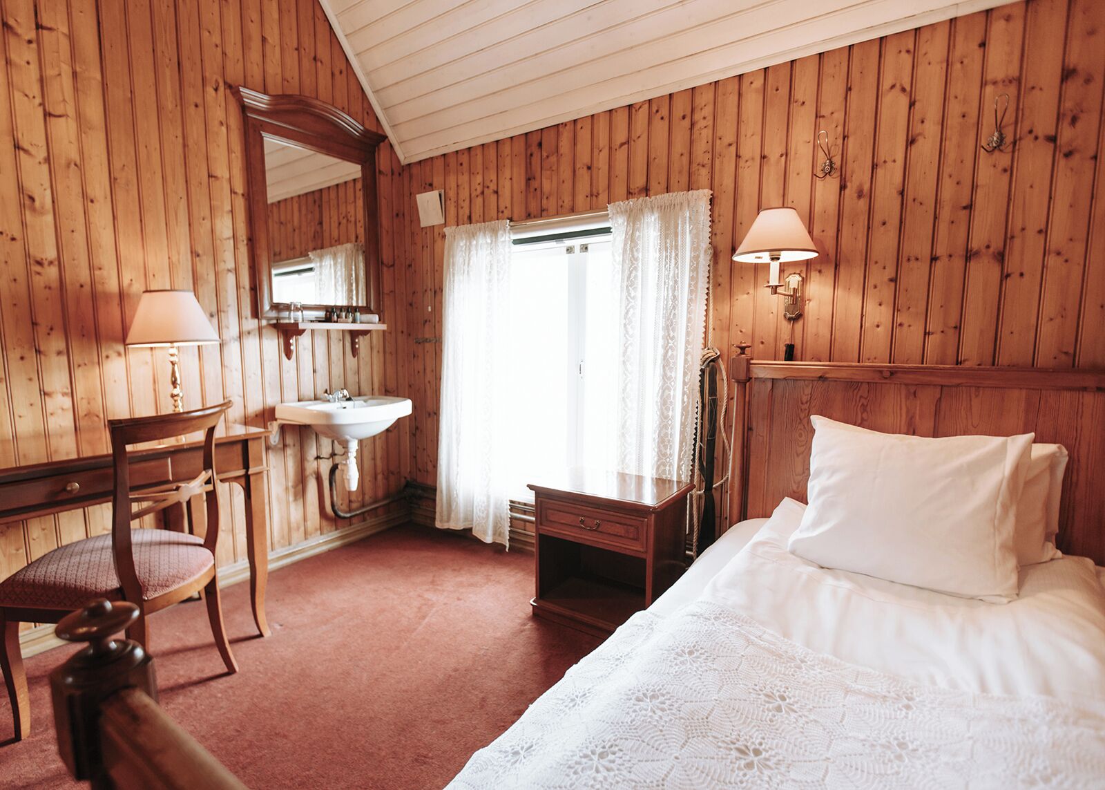 A wooden room with a bed, a desk, a chair, a sink, and a mirror.