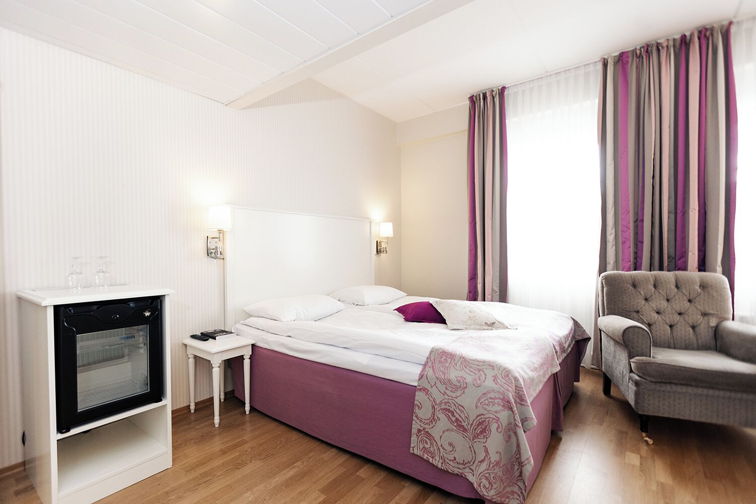 Una habitación de hotel con cama, una silla, una mesita y un minibar.