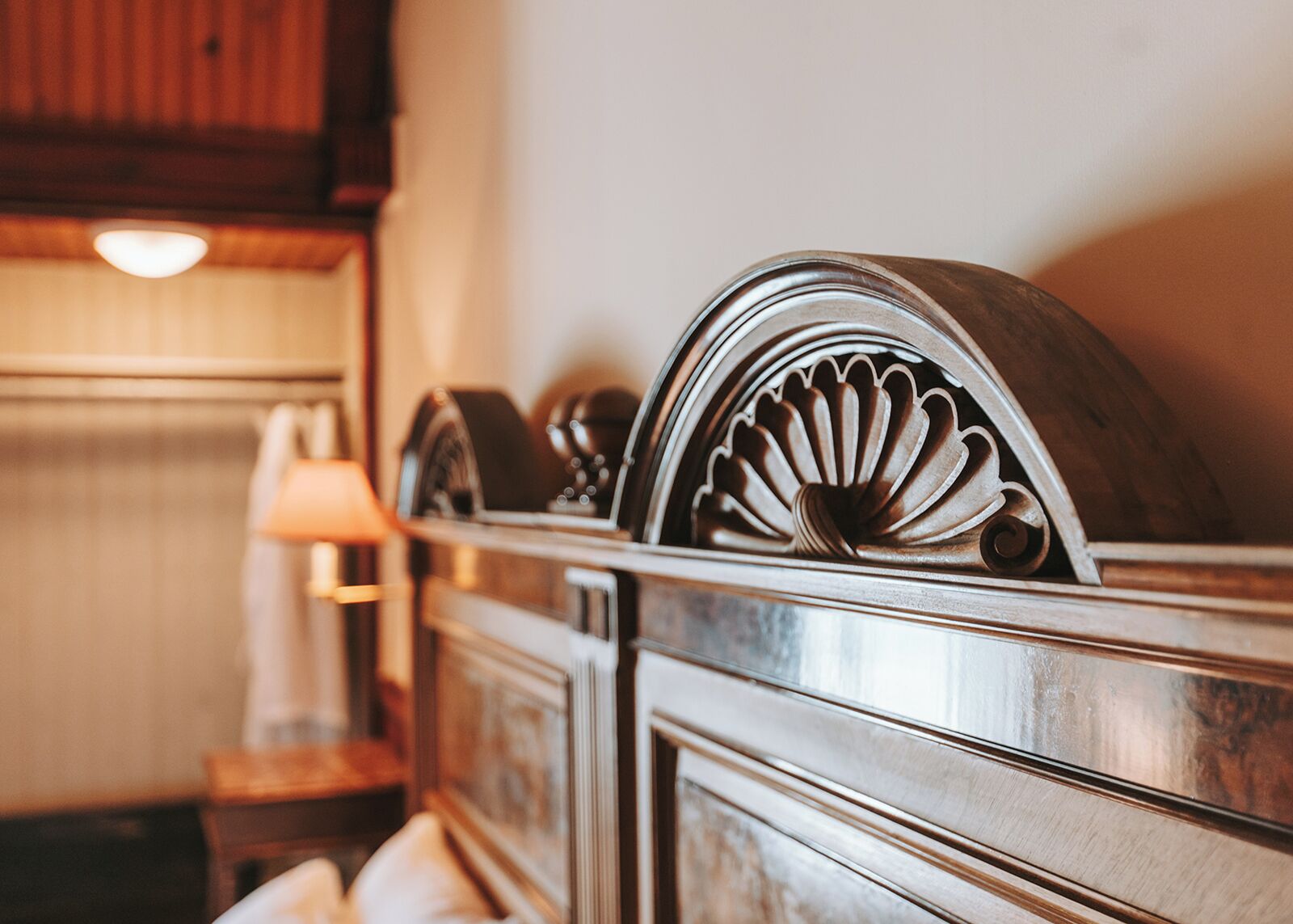 Een detail van een gepolijst houten hoofdbord met een decoratief metalen ornament, een lamp en een ingelijst beeld op een nachtkastje.