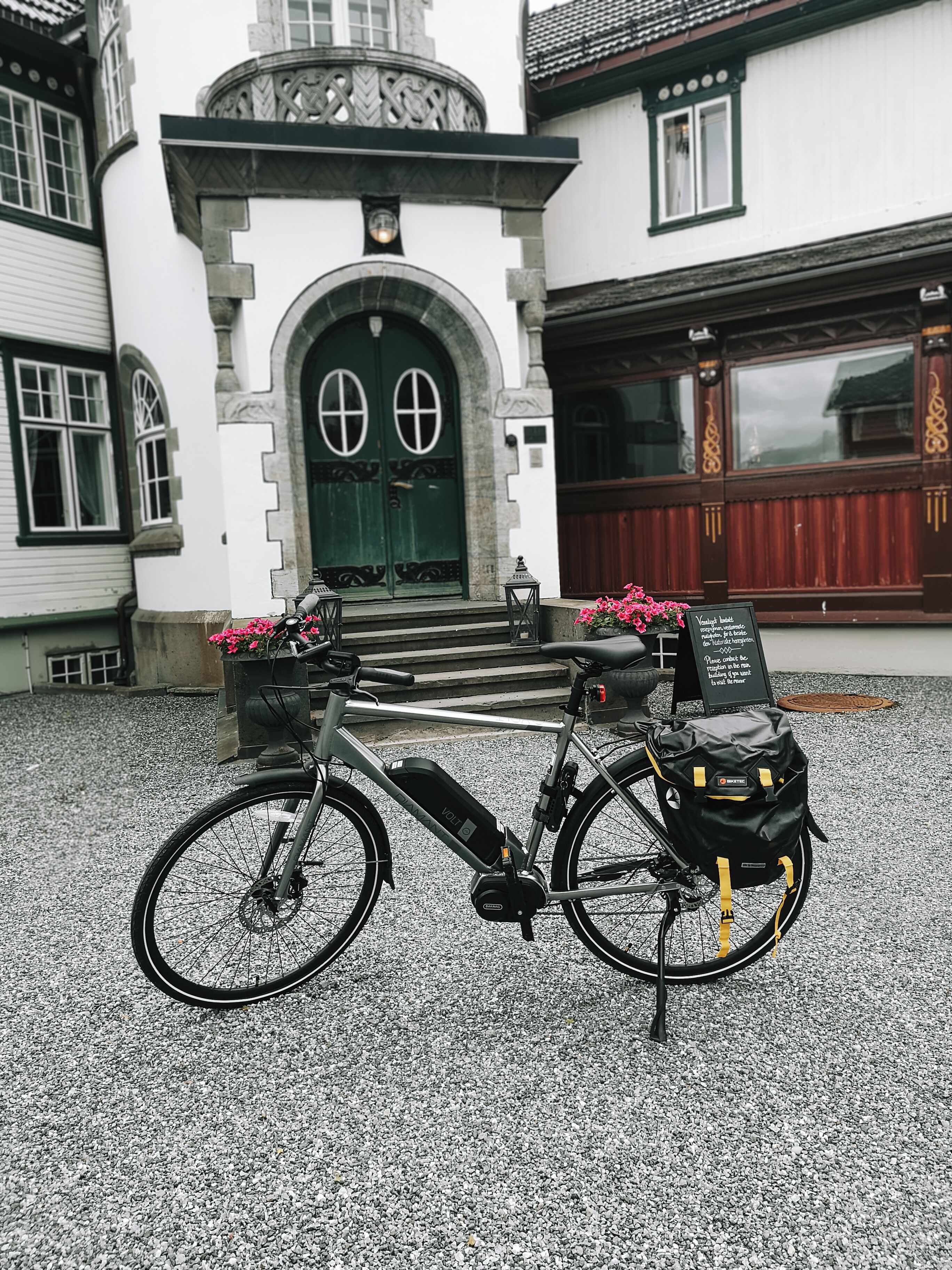 Un vélo d’appartement équipé d’un panier et d’un petit sac, garé sur une surface de gravier devant un bâtiment avec une porte verte et une enseigne.